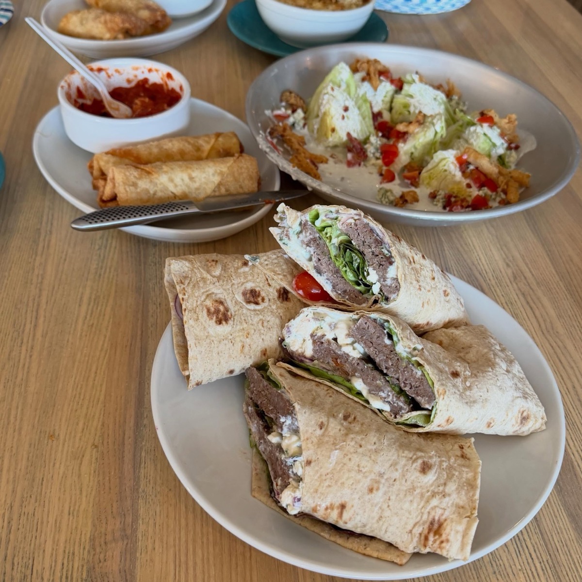 A spread of Market & Main dishes on a wood table: globally inspired wraps, a chopped salad, and crispy rolls